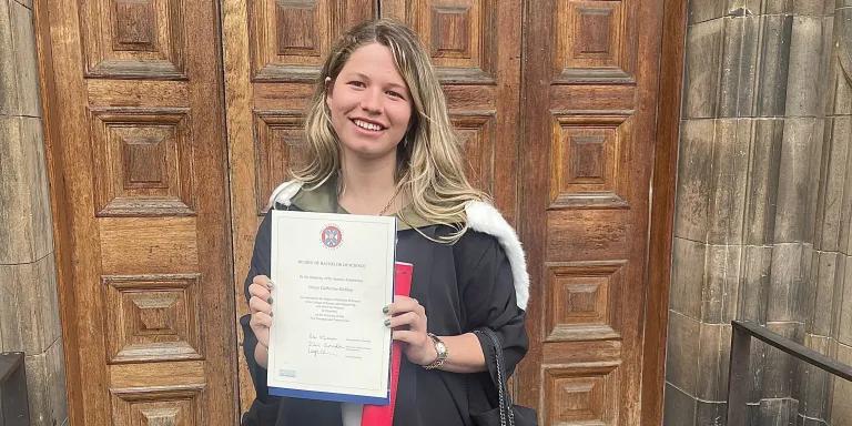 Alumna Grace Gubbay standing outside of the McEwan Hall with degree certificate