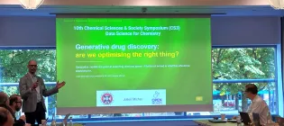 Professor Julien Michel is seen presenting in a small meeting room in from of a screen - slide title is "Generative drug discovery: are we optimising the right thing?"