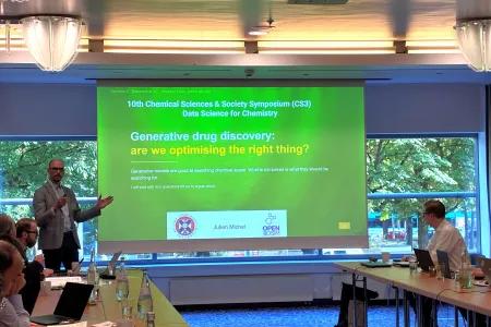 Professor Julien Michel is seen presenting in a small meeting room in from of a screen - slide title is "Generative drug discovery: are we optimising the right thing?"